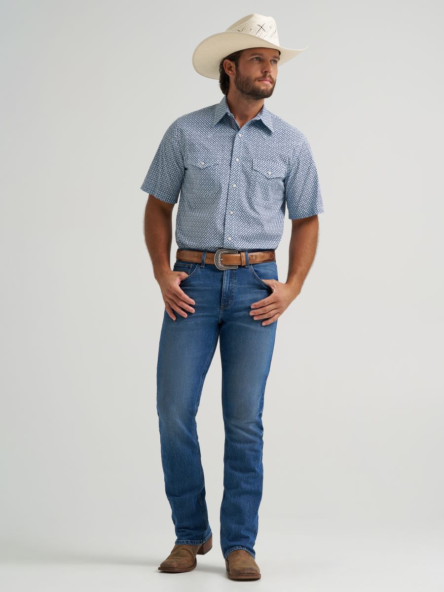 Man wearing a blue patterned Wrangler shirt, blue jeans, and a white cowboy hat on a light gray background