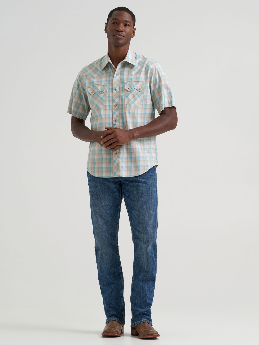 A man modeling a short-sleeve button-up Wrangler plaid shirt in light blue, white, and beige, paired with blue jeans and brown shoes, against a plain light gray studio background.
