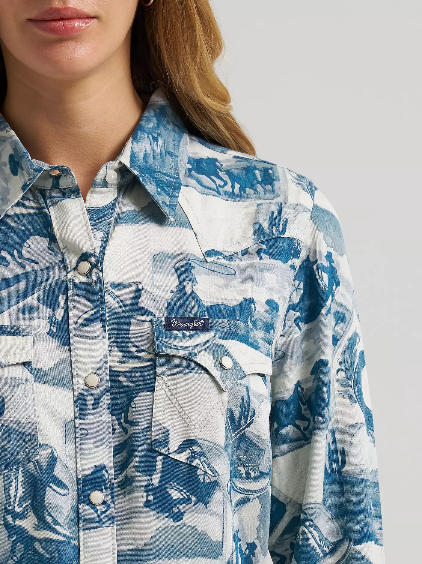 A close-up of a woman wearing a blue and white printed button-up shirt from Wrangler with a visible Wrangler logo patch on the chest pocket.