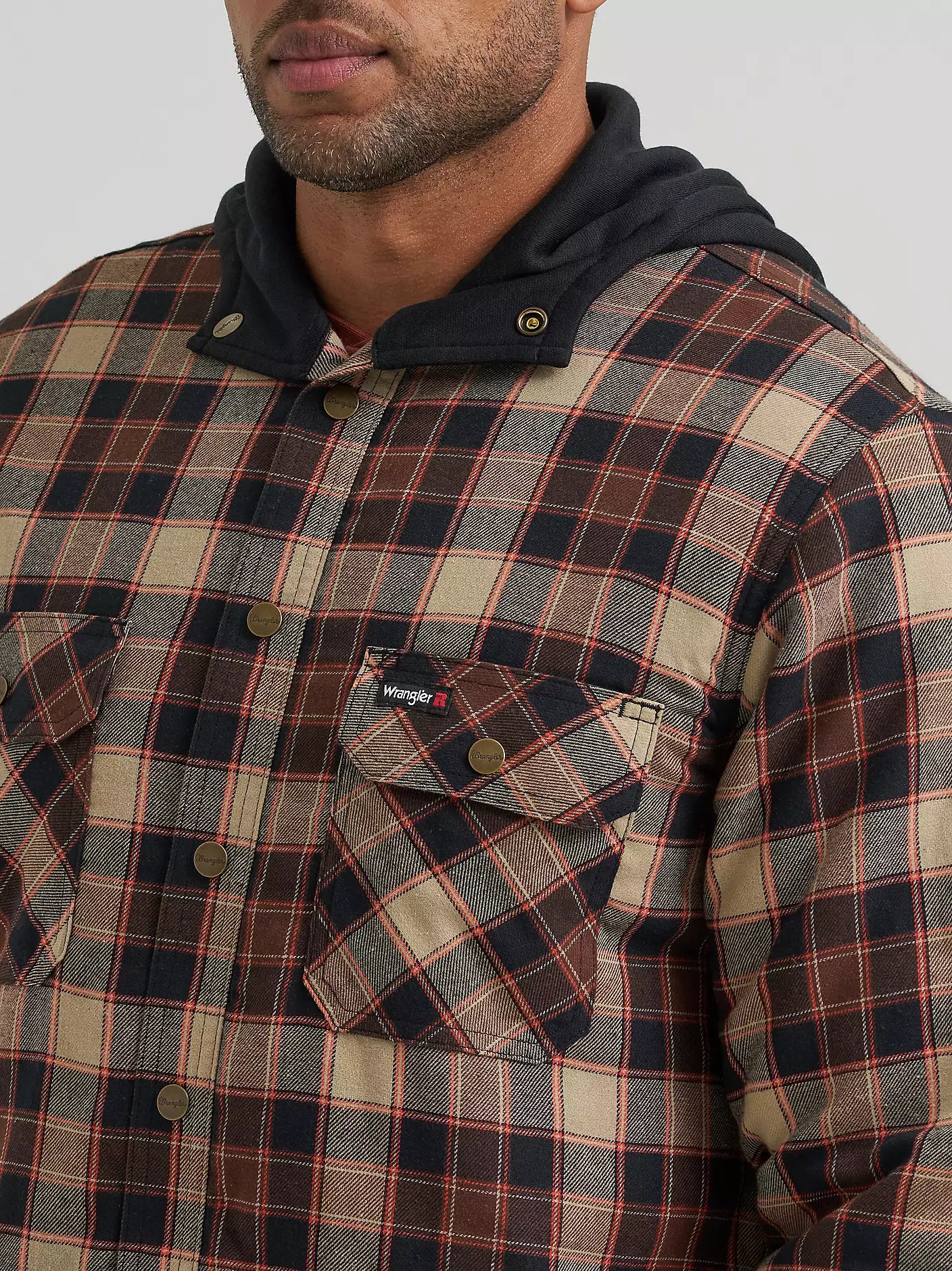 A close-up of a man wearing a plaid hooded Wrangler jacket. The garment features a beige, brown, and red plaid pattern with a dark navy hood and matching brown button details.
