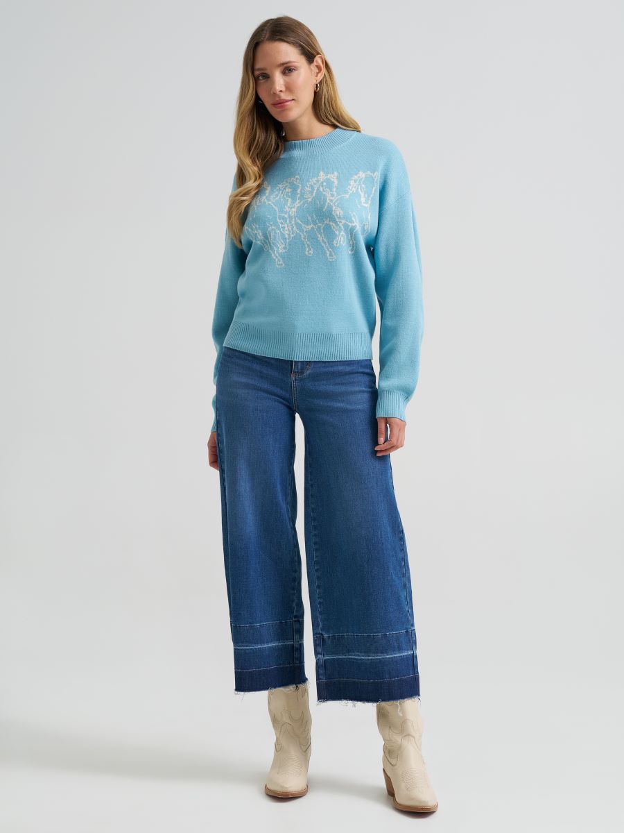 A woman modeling a light blue Wrangler crewneck sweater with a white embroidered mountain design, paired with wide-leg blue jeans and beige ankle boots, against a light gray studio background.