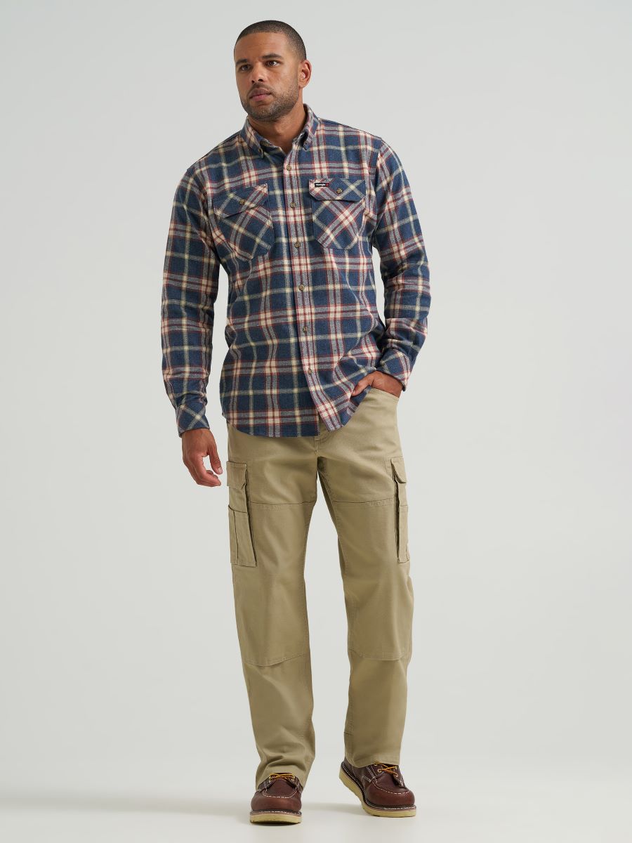 A man modeling a long-sleeve button-down plaid Wrangler shirt in navy, red, and beige, paired with khaki cargo pants and brown shoes, against a light gray studio background.