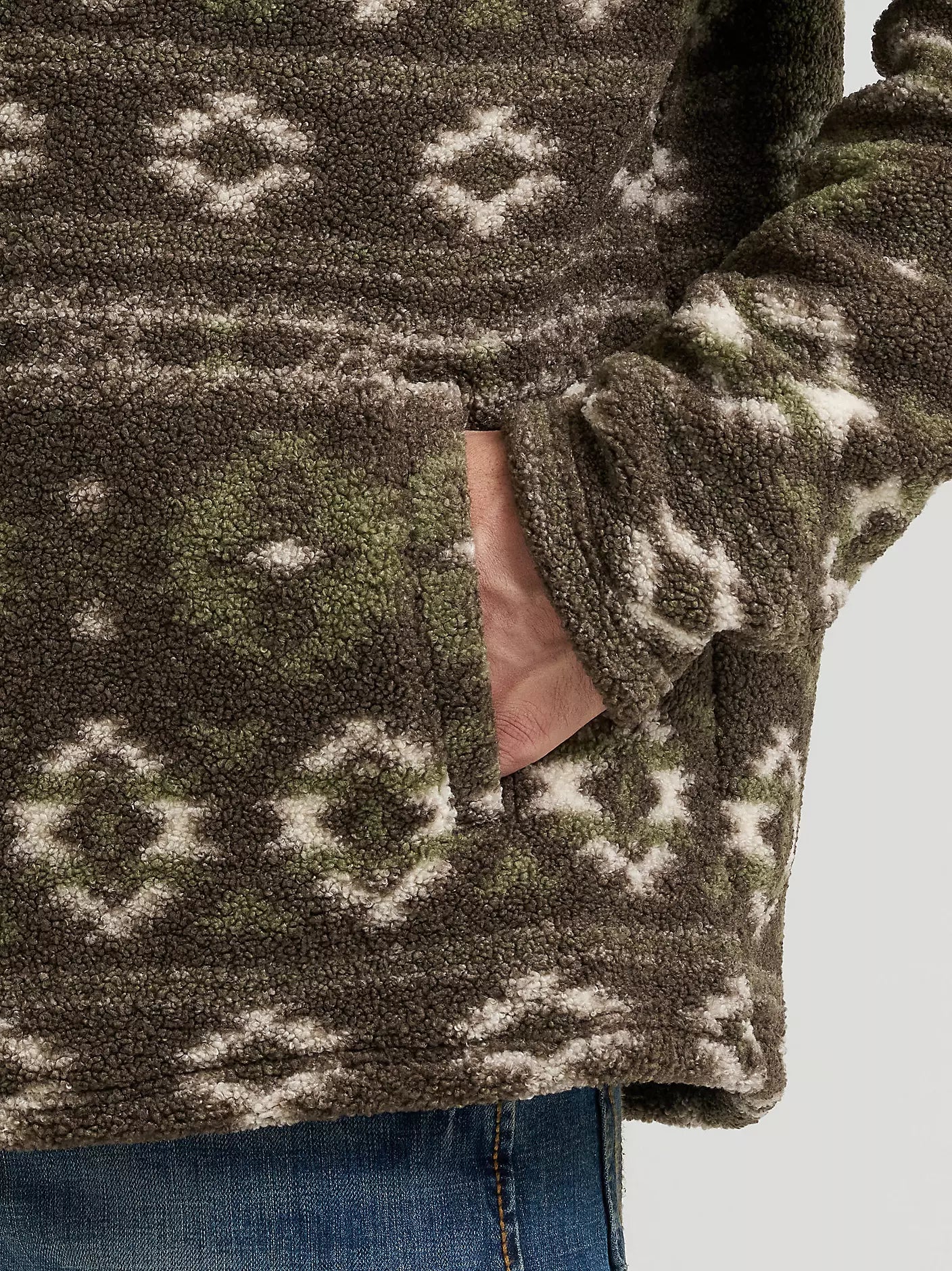 Close-up of a person wearing a textured fleece pullover from Wrangler with an earthy green and off-white geometric pattern, showing a hand in a pocket and blue jeans.