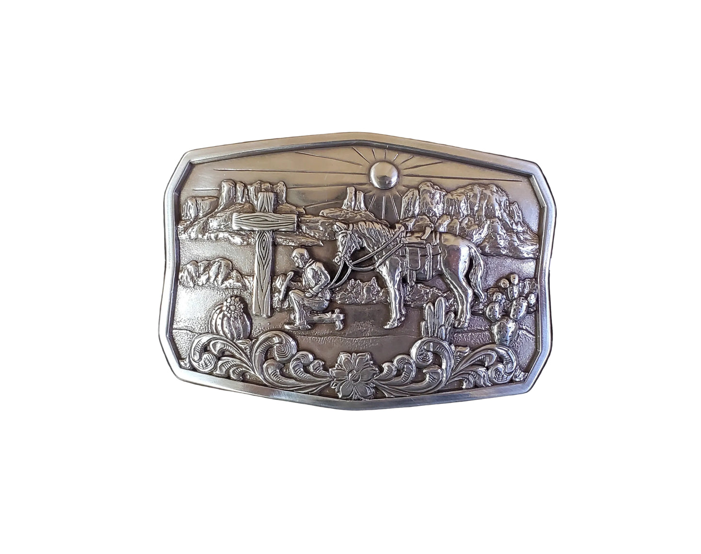 A Praying Desert Cowboy Belt Buckle by Nocona, displayed in its open box, showing a silver belt buckle with an engraved scene of a cowboy and a horse in a desert landscape.