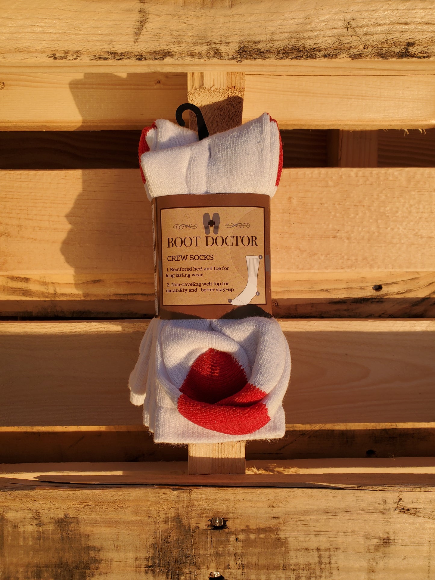 A pair of white crew height youth socks with a red design, hanging on a wooden surface with a label attached that reads 'Boot Doctor'.