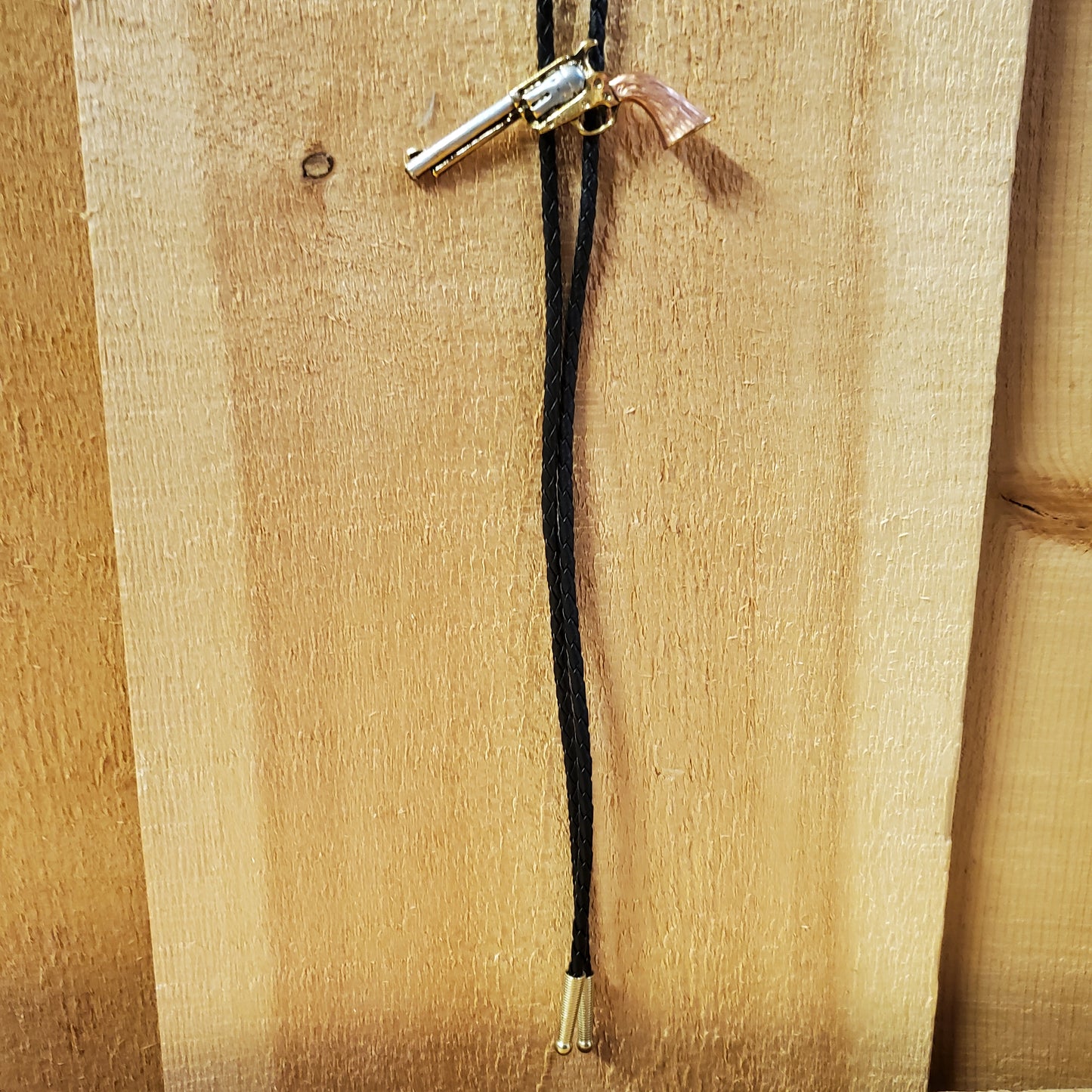 A tri-tone revolver bolo tie with a silver body and a brown barrel, attached to a black rope and a wooden surface.