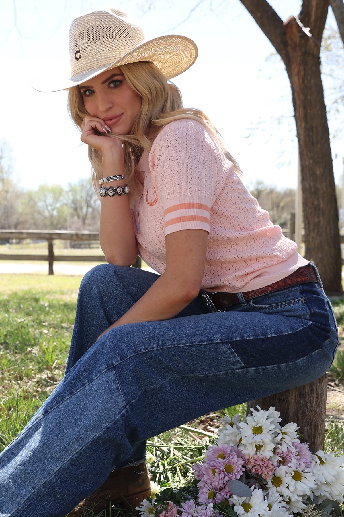 Woman in a pink shirt and blue jeans sitting outdoors with flowers and a hat.