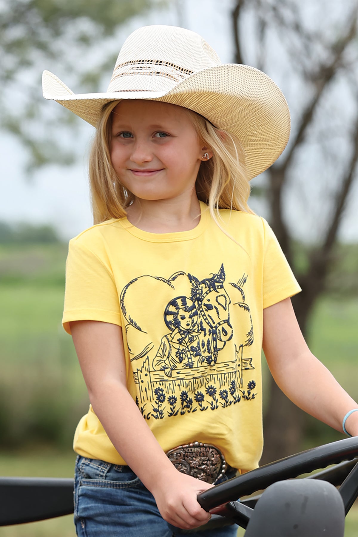 Child wearing a yellow Cruel Girls t-shirt with a horse graphic and a cowboy hat, standing outdoors.