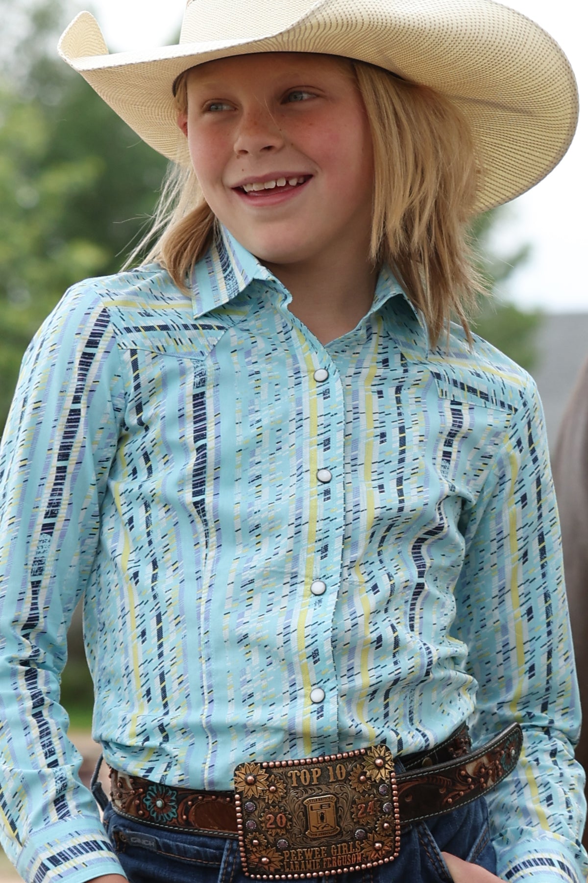 Child wearing a light blue patterned Cinch shirt with a cowboy hat outdoors.