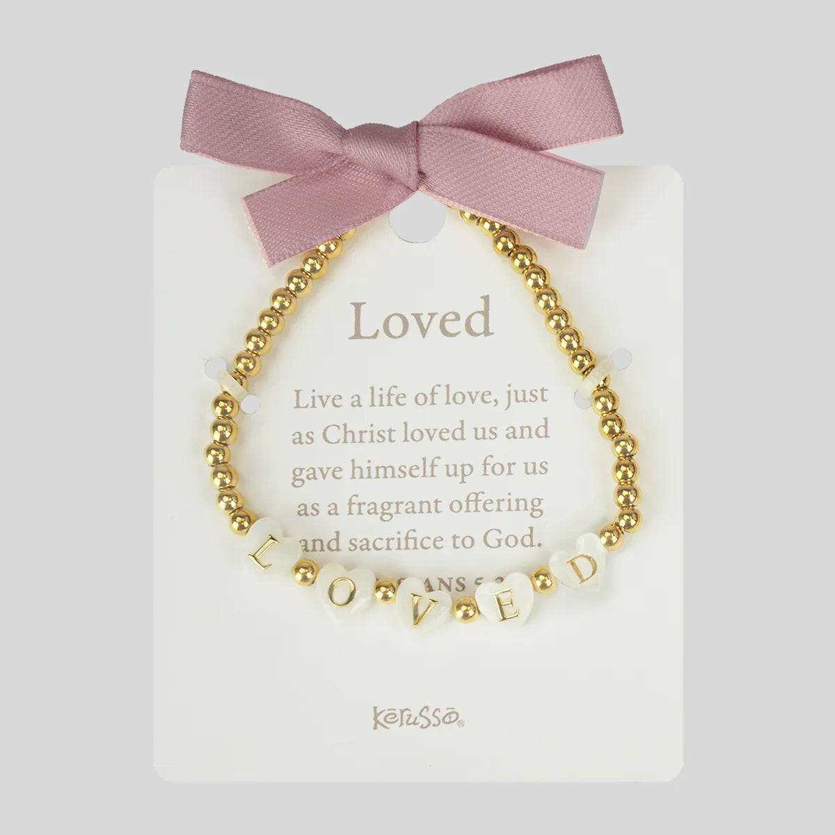 A gold beaded bracelet with white letter beads spelling 'LOVED' and small gold beads, displayed on a white card with a pink ribbon bow. The card reads 'Loved' with a Bible verse and the brand name 'Kerusso' at the bottom.