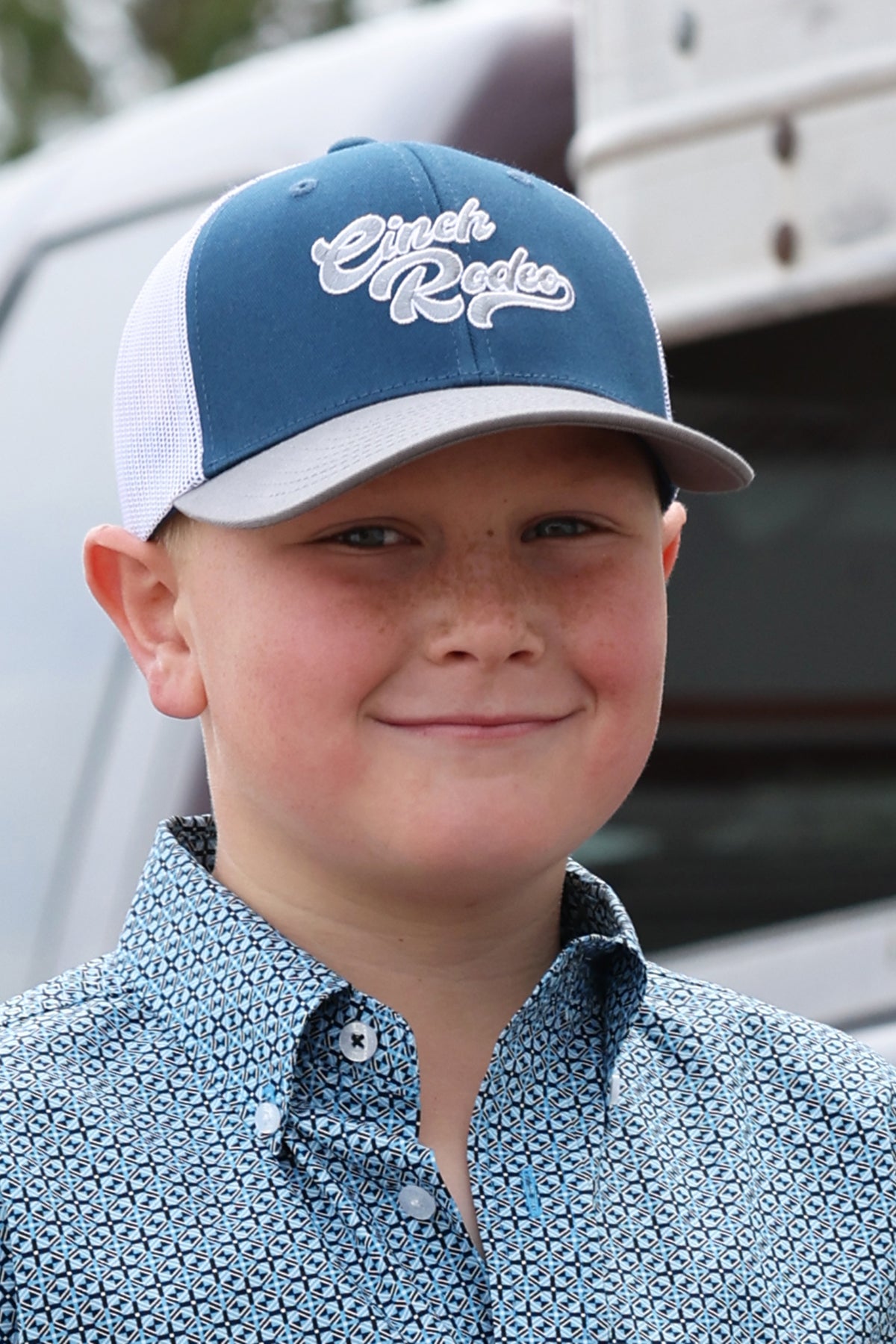 Young boy wearing a blue and white Cinch ball cap with text, standing outdoors.