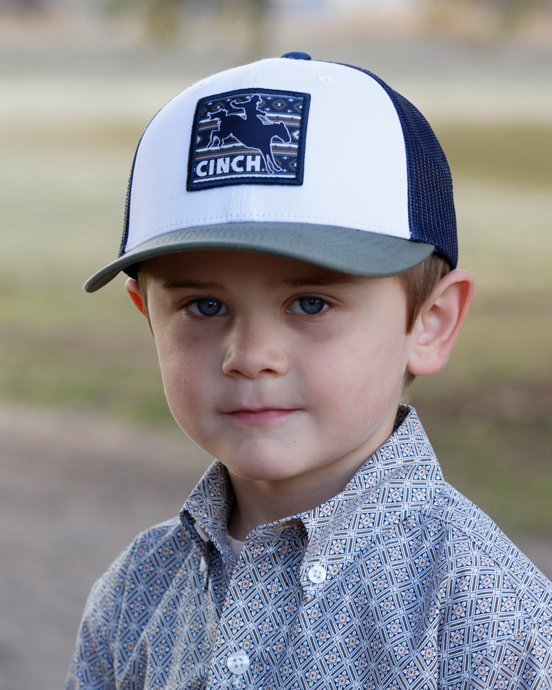 Child wearing a white and navy blue Cinch cap with a logo, standing outdoors.