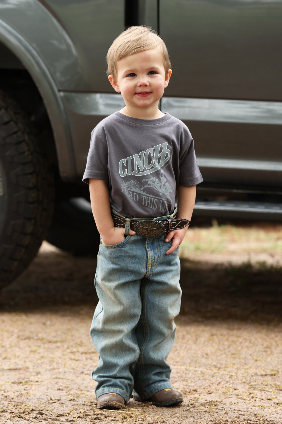 Child wearing a gray Cinch t-shirt and blue jeans standing in front of a vehicle.