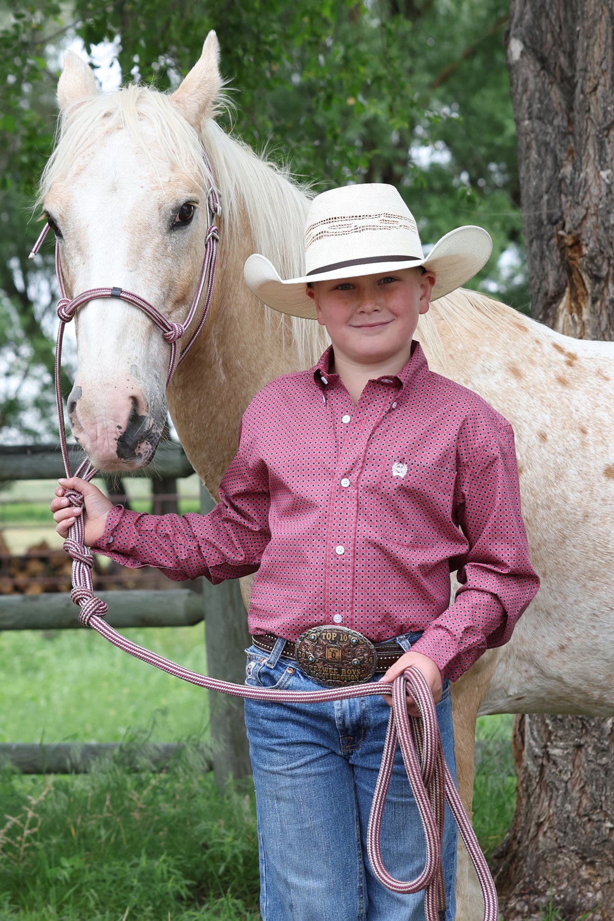 Person in cowboy hat and Cinchshirt standing next to a horse with a rope, outdoors.