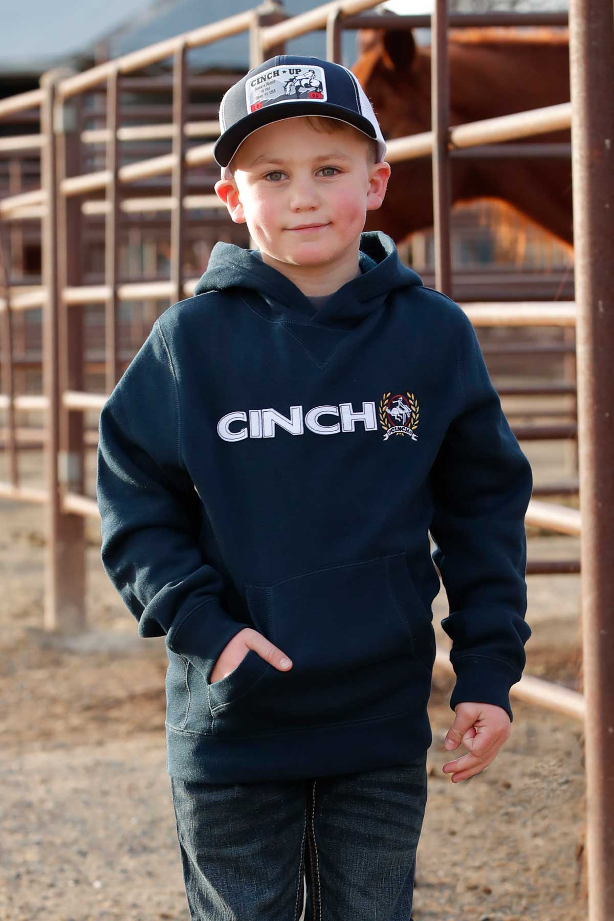Boy wearing a navy Cinch sweater, blue jeans and ball cap walking in a horse farm