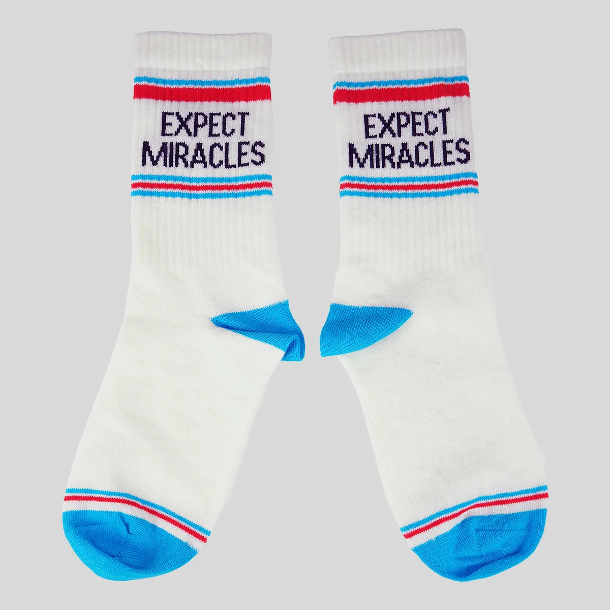 A pair of white crew socks from Kerusso with blue and red stripes, featuring the text 'EXPECT MIRACLES' in navy blue on the upper cuff, and bright blue heels and toes, set against a solid gray background.