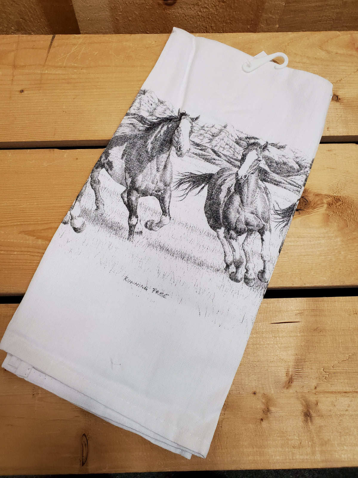 A white cotton tea towel with a printed black and gray running horse design, laying on a wooden surface.