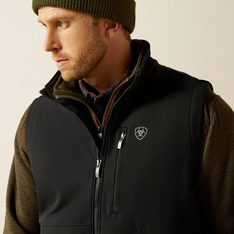 Man wearing a black Ariat vest, brown sweater,  and a green toque on a cream colored background