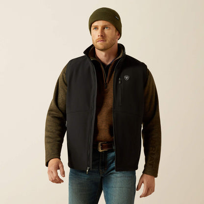 Man wearing a black Ariat vest, brown sweater, blue jeans and a green toque on a cream colored background