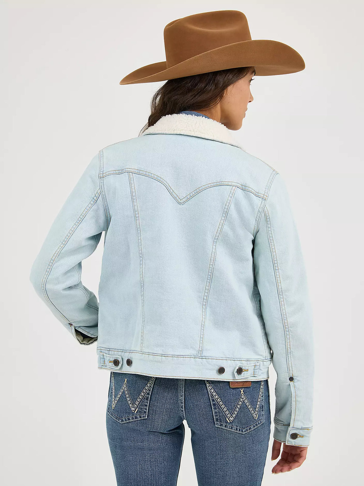 Woman wearing a light denim Wrangler sherpa lined jacket, blue jeans and a brown cowboy hat on a white background