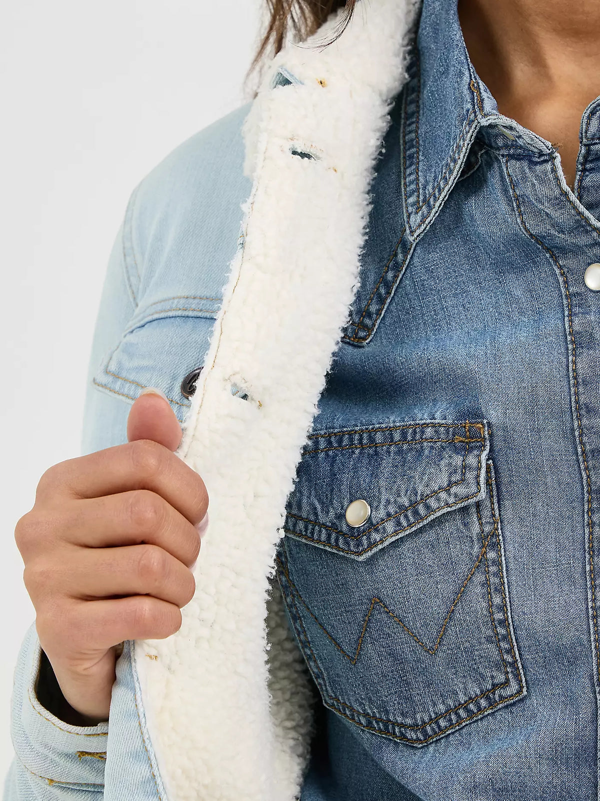 Woman wearing a light denim Wrangler sherpa lined jacket, and a denim shirt on a white background