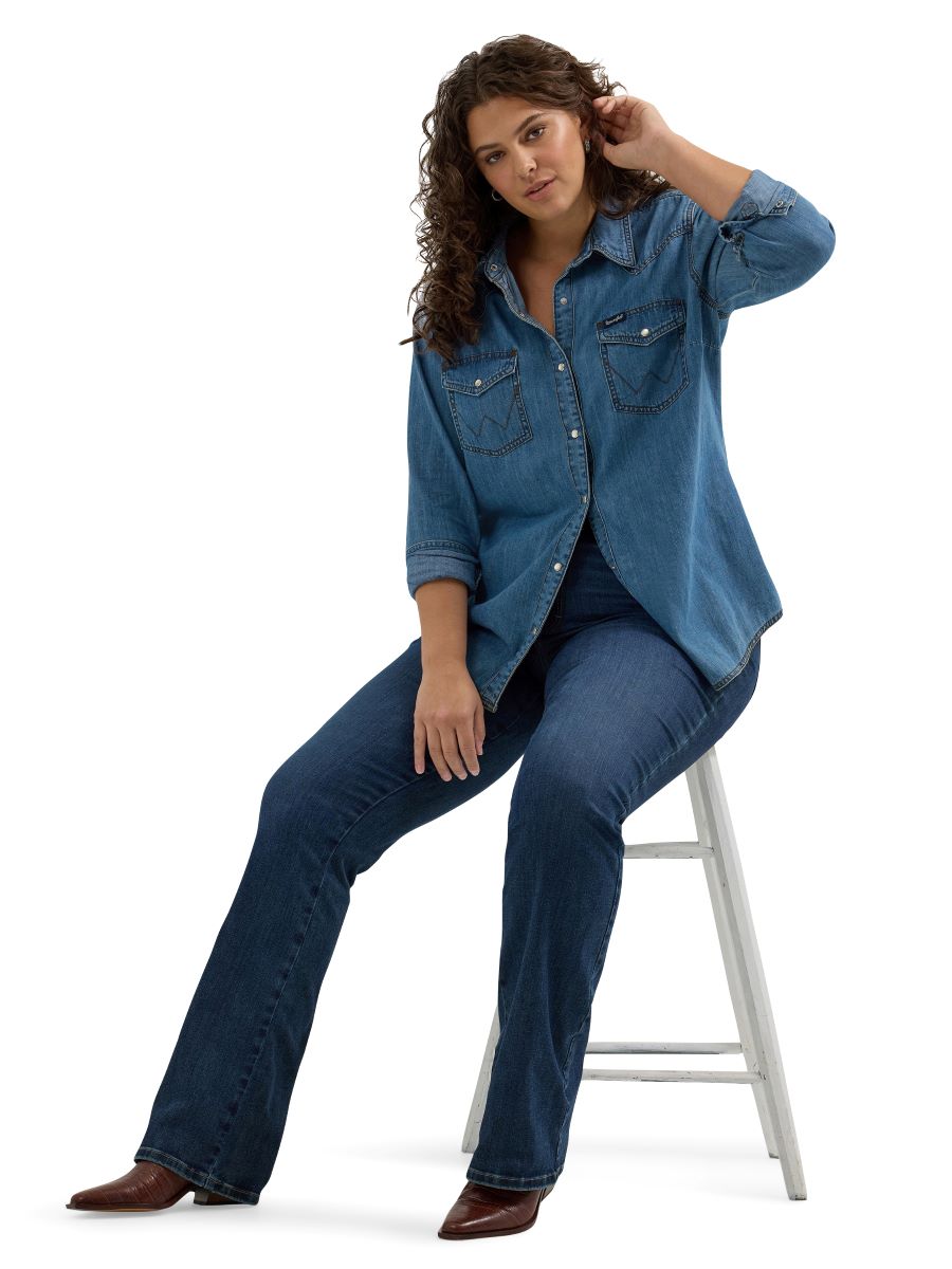 Woman wearing a blue denim shirt and jeans sitting on a stool against a white background
