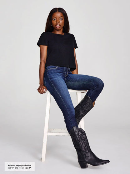 Woman wearing blue jeans and black cowboy boots sitting on a white stool against a light gray background.