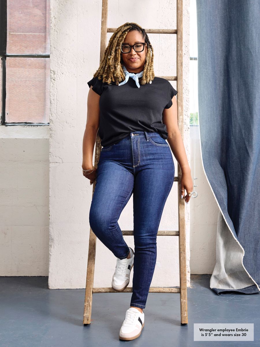 Woman in black top and blue jeans standing on a ladder in a room with a window and curtain.