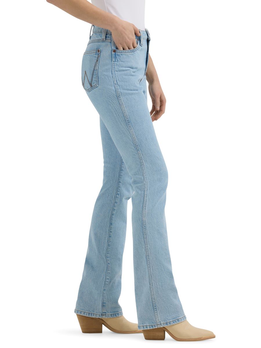 A side view of a woman wearing light wash 'Bailey' high rise boot cut women's jeans by Wrangler, with a leather patch on the pocket and a button closure.