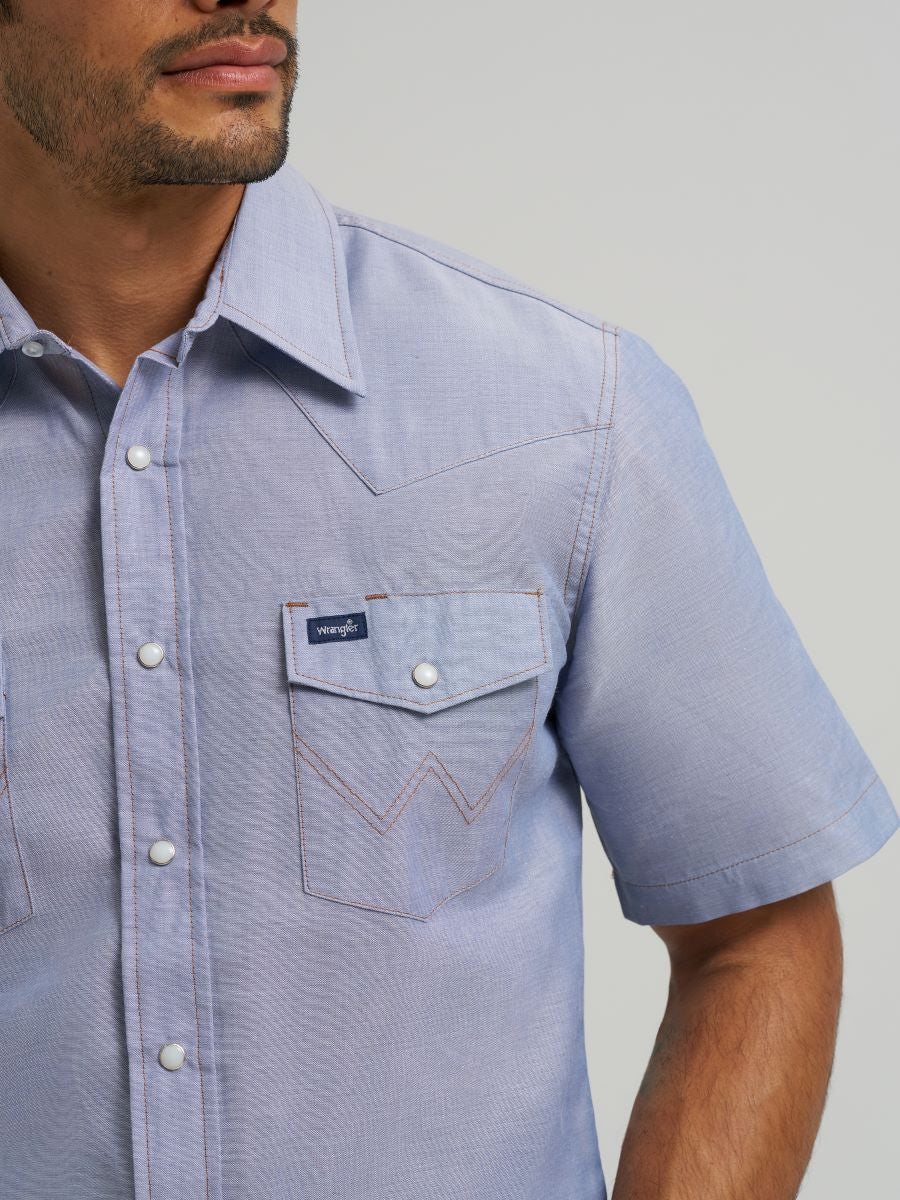 Man wearing a light blue short-sleeve Wrangler shirt with a brand logo on a plain background