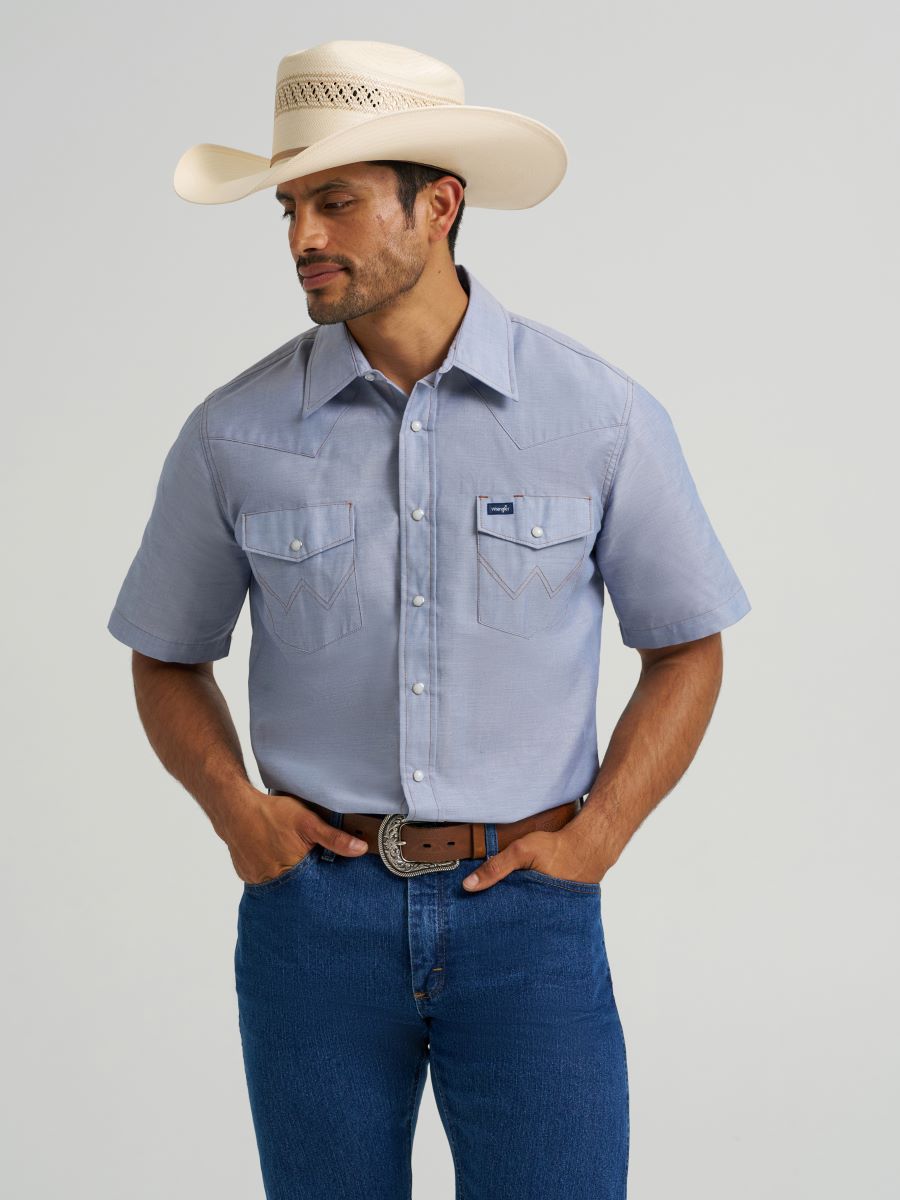 Man wearing a light blue short-sleeve Wrangler shirt, blue jeans, and a cowboy hat on a gray background