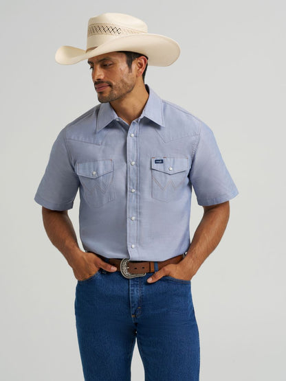 Man wearing a light blue short-sleeve Wrangler shirt, blue jeans, and a cowboy hat on a gray background
