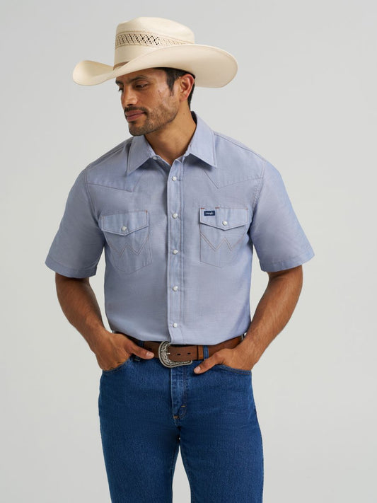 Man wearing a light blue short-sleeve Wrangler shirt, blue jeans, and a cowboy hat on a gray background