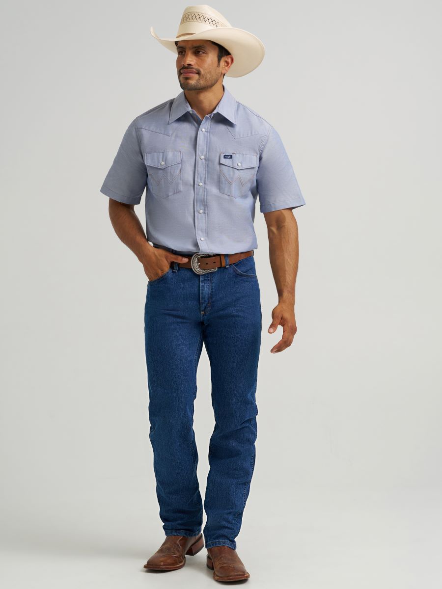 A man modeling a light blue short-sleeve Wrangler western button-down shirt with two chest pockets, paired with dark blue jeans, a brown belt, and a cream cowboy hat against a light gray studio background.