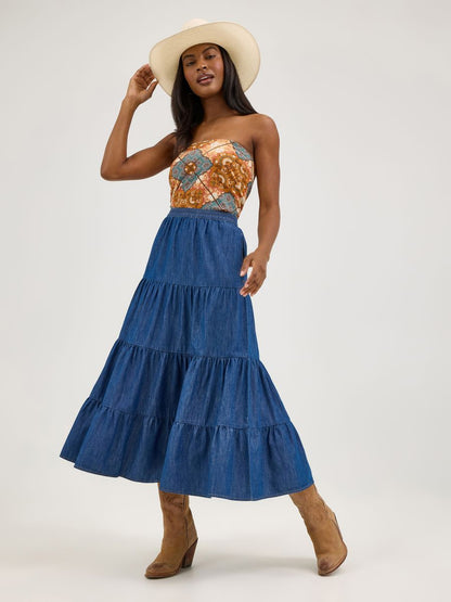 Woman wearing a strapless floral top and blue tiered denim skirt with a hat on a white background
