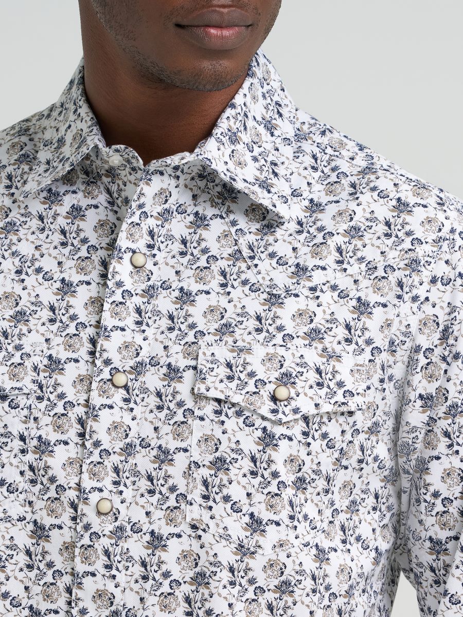 Man wearing a patterned Wrangler shirt, blue jeans, and a dark cowboy hat on a light gray background