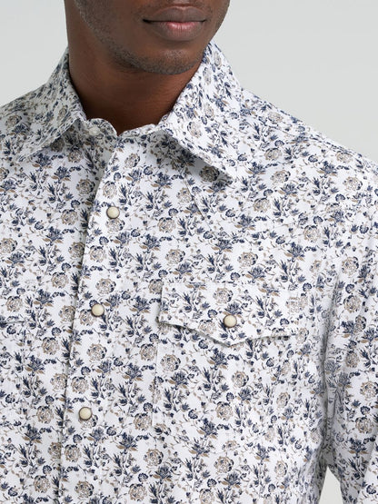 Man wearing a patterned Wrangler shirt, blue jeans, and a dark cowboy hat on a light gray background