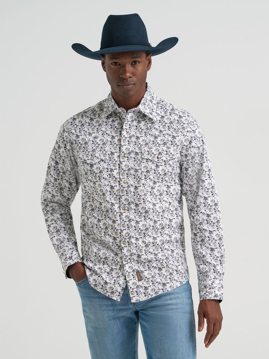 Man wearing a patterned Wrangler shirt, blue jeans, and a dark cowboy hat on a light gray background