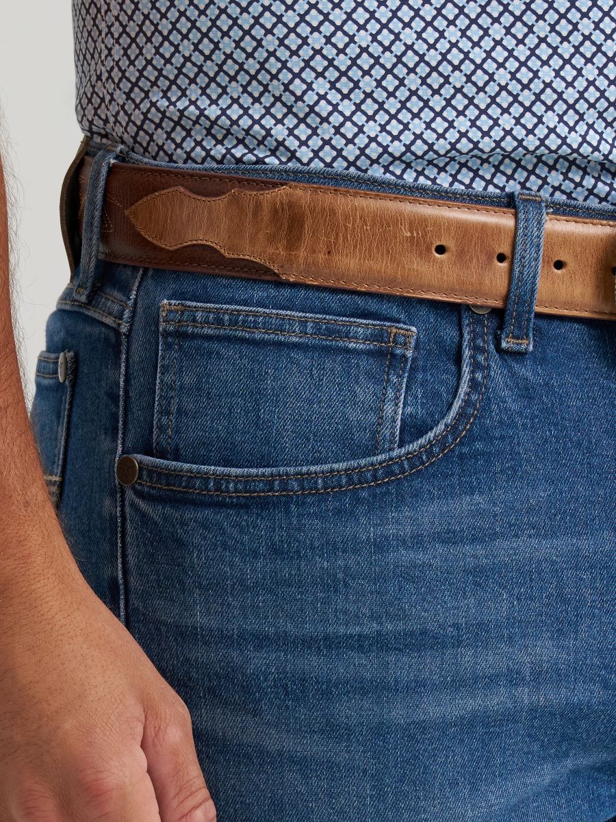 A close-up of a person wearing a brown leather belt with a decorative stitched center, paired with blue Wranlger jeans and a patterned shirt.