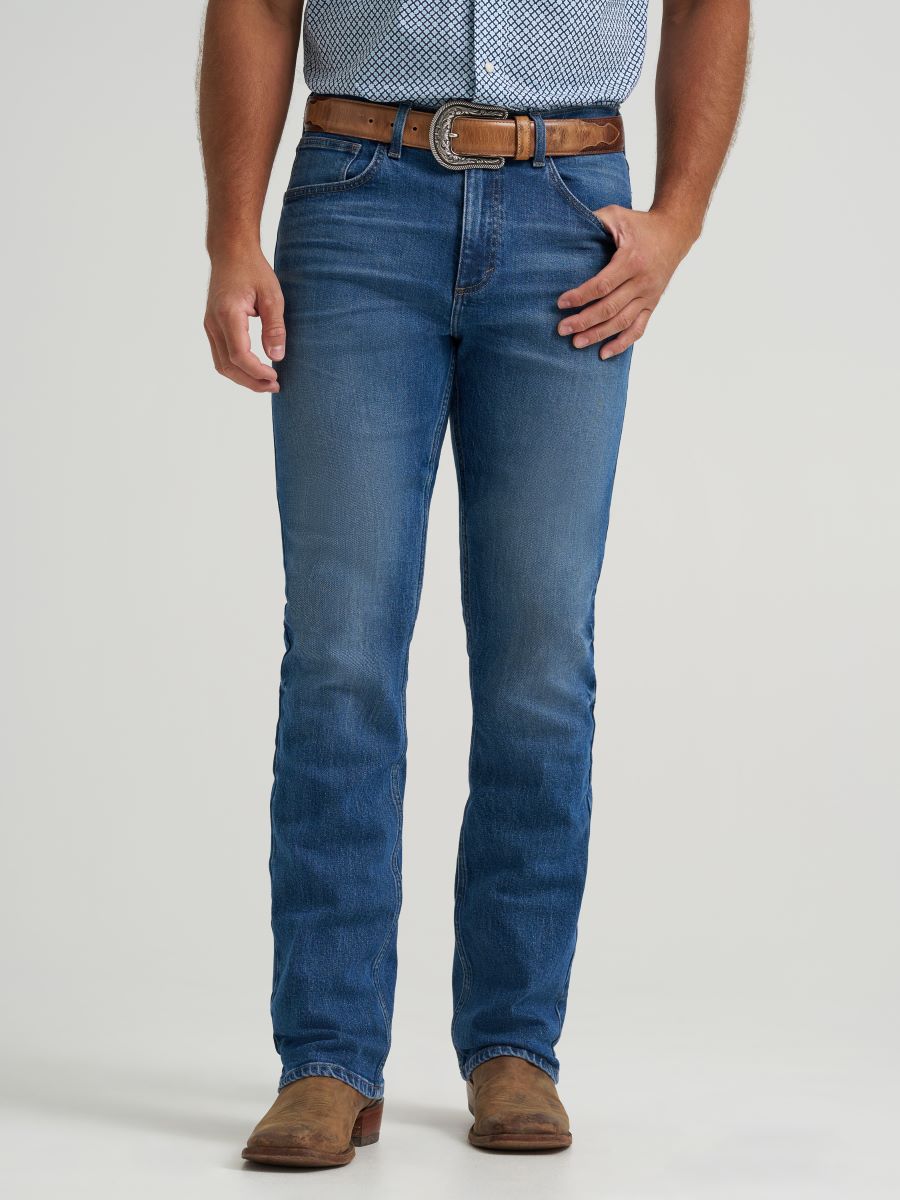 man wearing blue denim Wrangler jeans with brown leather belt and brown shoes, against a light gray studio background.