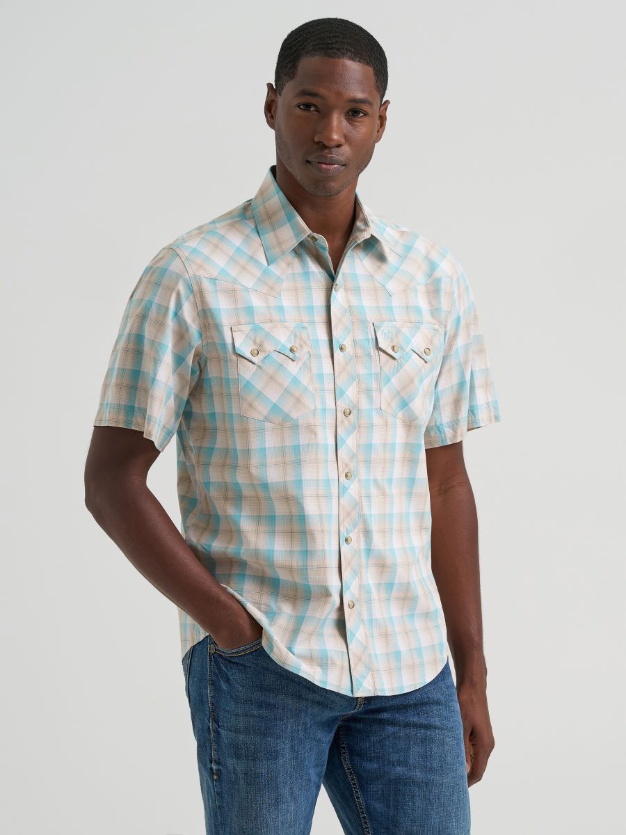 A man wearing a short-sleeve button-up Wrangler plaid shirt in a light peach, cream, and light blue colorway, paired with blue jeans, against a solid light gray background.