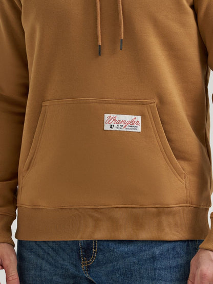 Close-up of a person wearing a tan-brown Wrangler pullover hoodie with a front kangaroo pocket and a visible Wrangler label on the pocket. Blue jeans are also visible at the bottom.