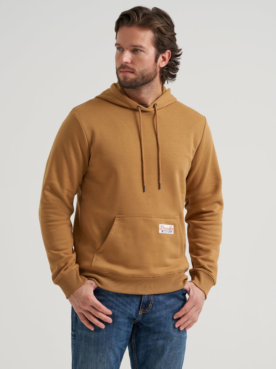 A man wearing a camel brown pullover hoodie by Wrangler with a drawstring hood and a front kangaroo pocket, paired with blue jeans, photographed against a light gray studio background.