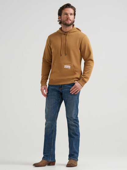 A man wearing a camel brown Wrangler pullover hoodie with a small front patch, blue jeans, and brown boots, standing against a light gray studio background.