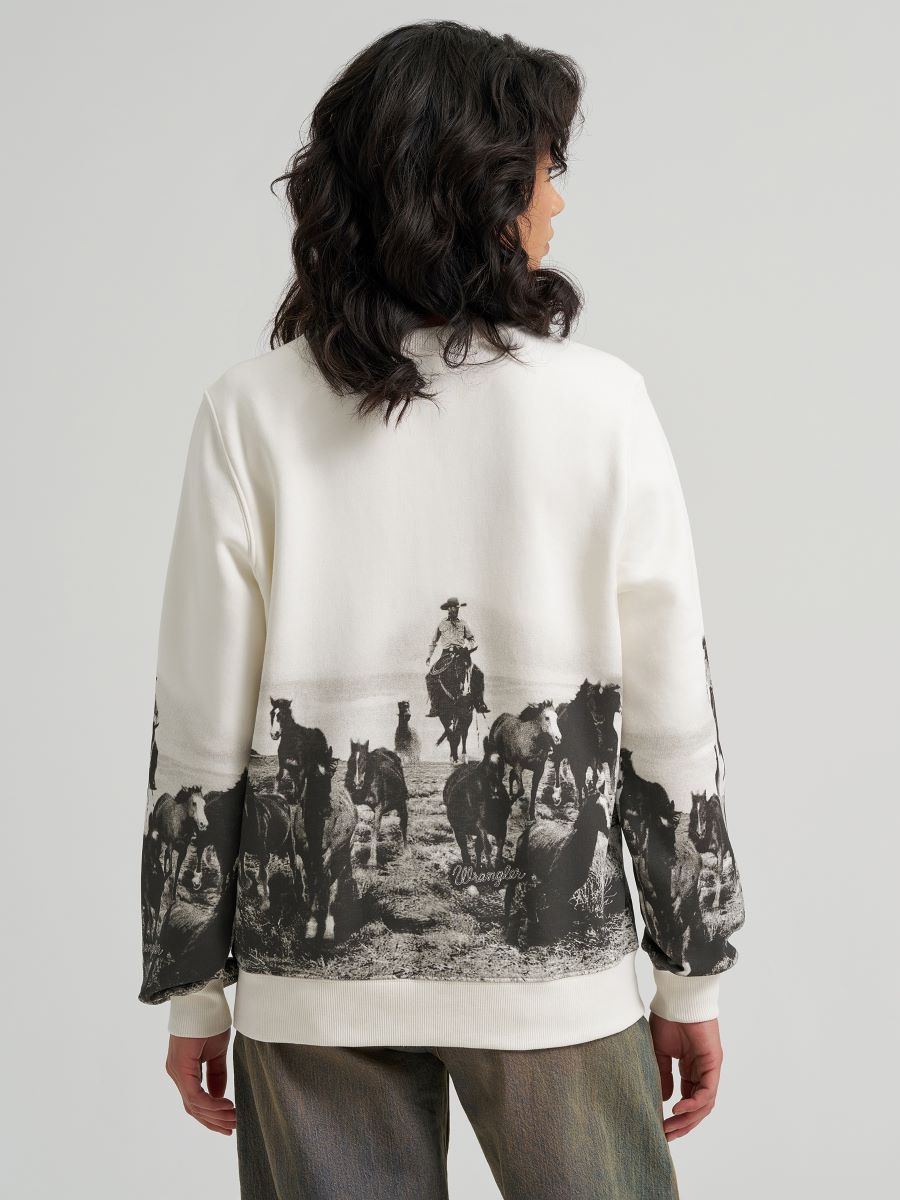 A model shown from the back wearing a cream-colored Wrangler sweatshirt with a large black-and-white western ranch print on the back and sleeves, paired with gray pants, against a light gray studio background.