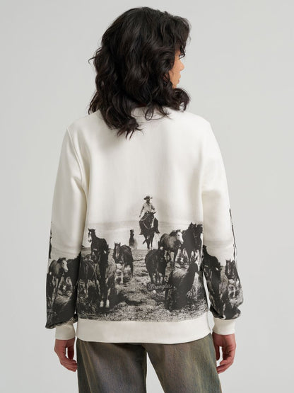 A model shown from the back wearing a cream-colored Wrangler sweatshirt with a large black-and-white western ranch print on the back and sleeves, paired with gray pants, against a light gray studio background.