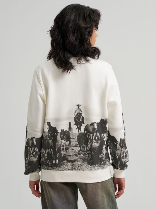 A model shown from the back wearing a cream-colored Wrangler sweatshirt with a large black-and-white western ranch print on the back and sleeves, paired with gray pants, against a light gray studio background.