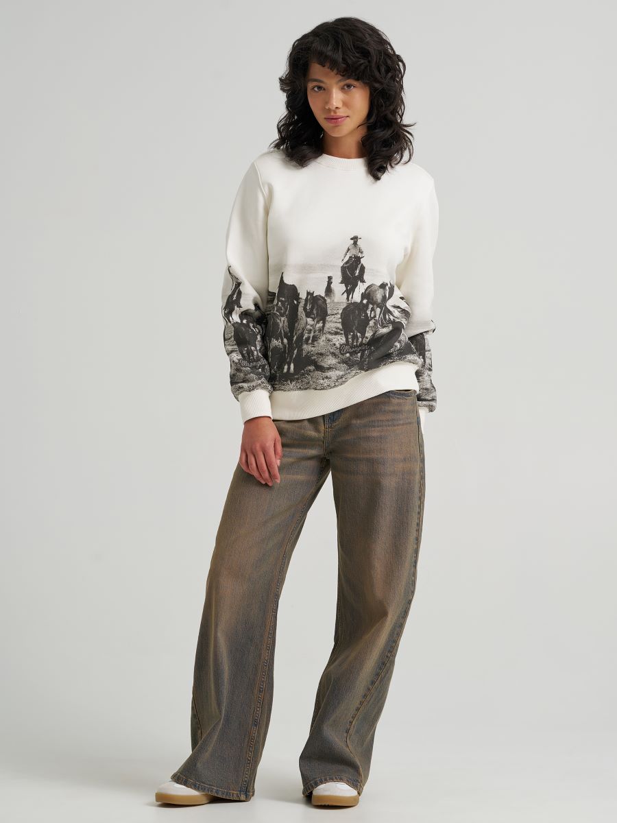 Woman wearing a white Wrangler sweater with black graphic design and brown pants on a light gray background