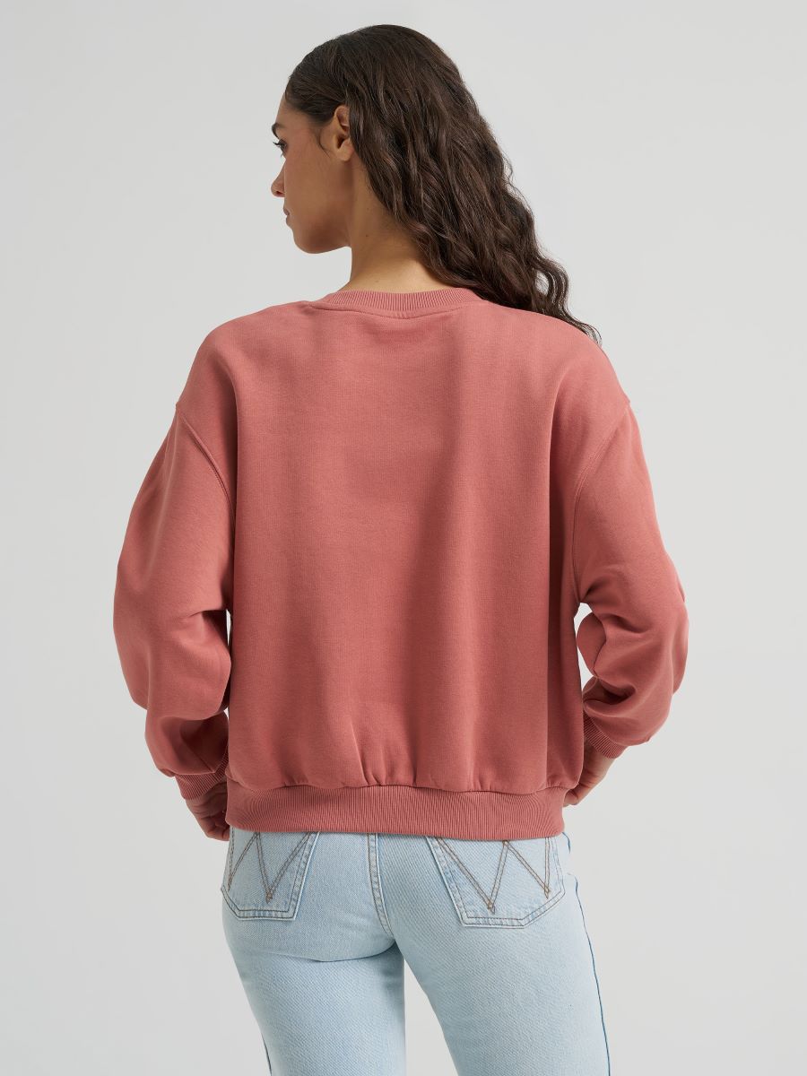 A woman shown from the back wearing a dusty rose Wrangler crewneck sweatshirt and light blue jeans against a plain light gray studio background.