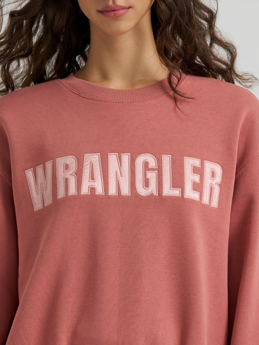 A close-up of a model wearing a dusty rose crewneck sweatshirt with 'WRANGLER' printed across the chest in a matching pink arched block font.